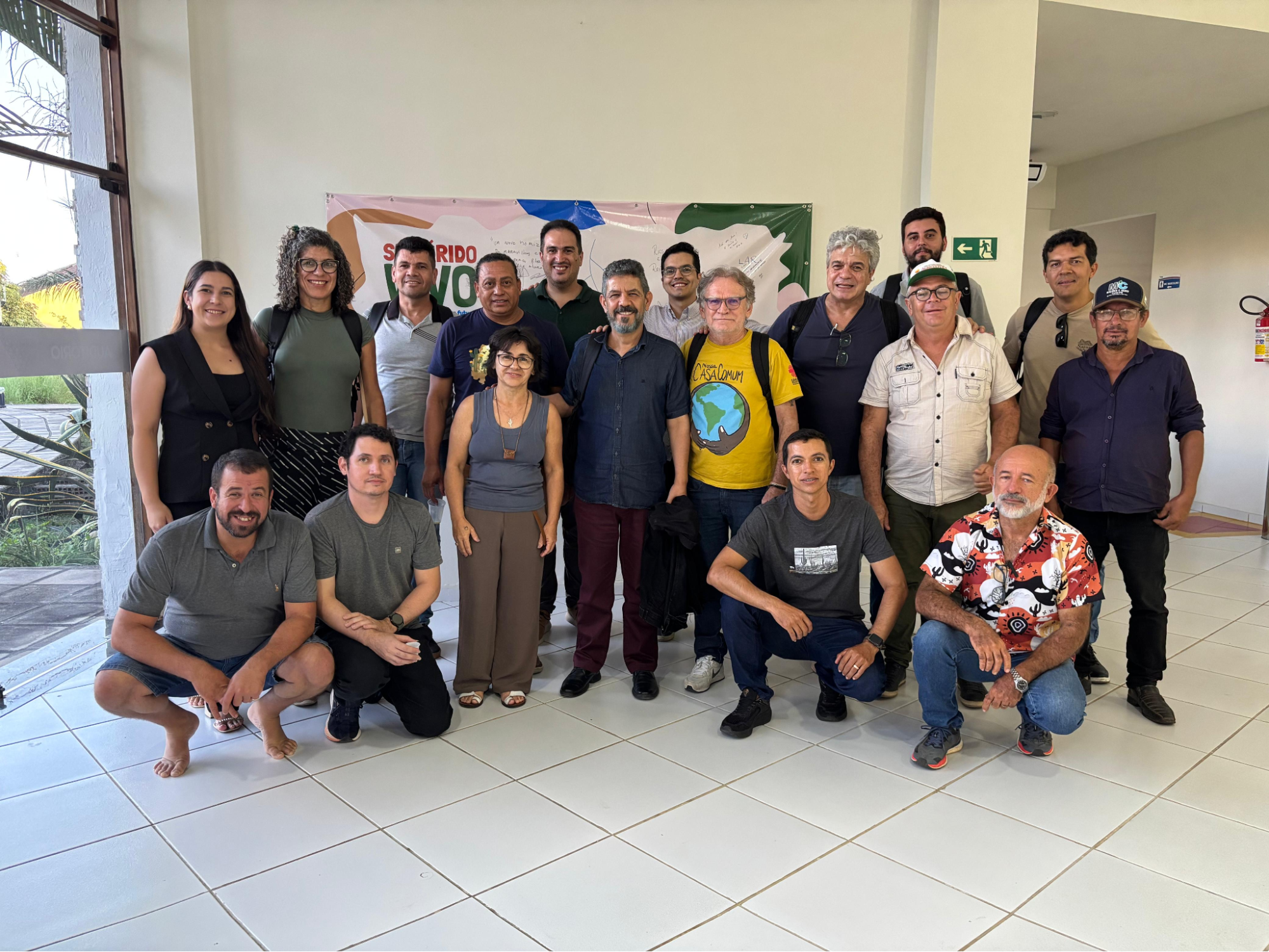 Fotografia de grupo com 17 pessoas reunidas em um ambiente interno, algumas em pé e outras agachadas à frente, posando e sorrindo para a câmera. Ao fundo, há um painel colorido com a inscrição “Semiárido Vivo”.