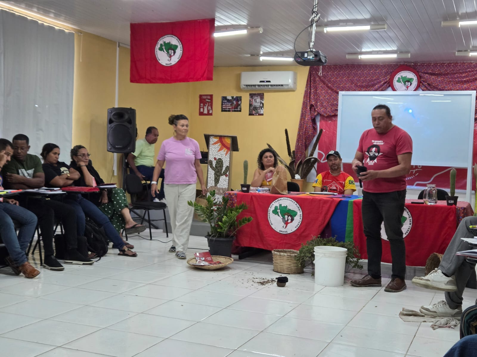 Atividade em sala com participantes sentados e duas pessoas em pé. Ao fundo, mesa e bandeiras vermelhas com o símbolo do MST.