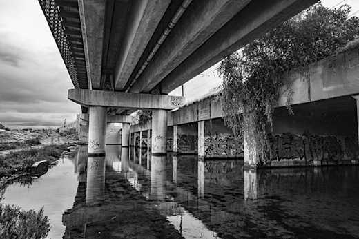 Black and white photograph of the reflections of a concrete overpass strewn with graffiti, an abandoned and forgotten space.