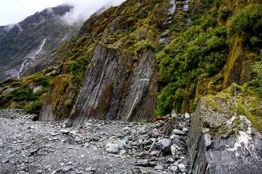 Photograph of dark rock face covered in lush vegetation as it meets the moraine covered surface of the glacial river bed.