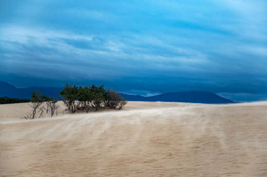 Photograph of sand dune in inclement weather with sand being whipped up and ominous dark mountains in the background. 