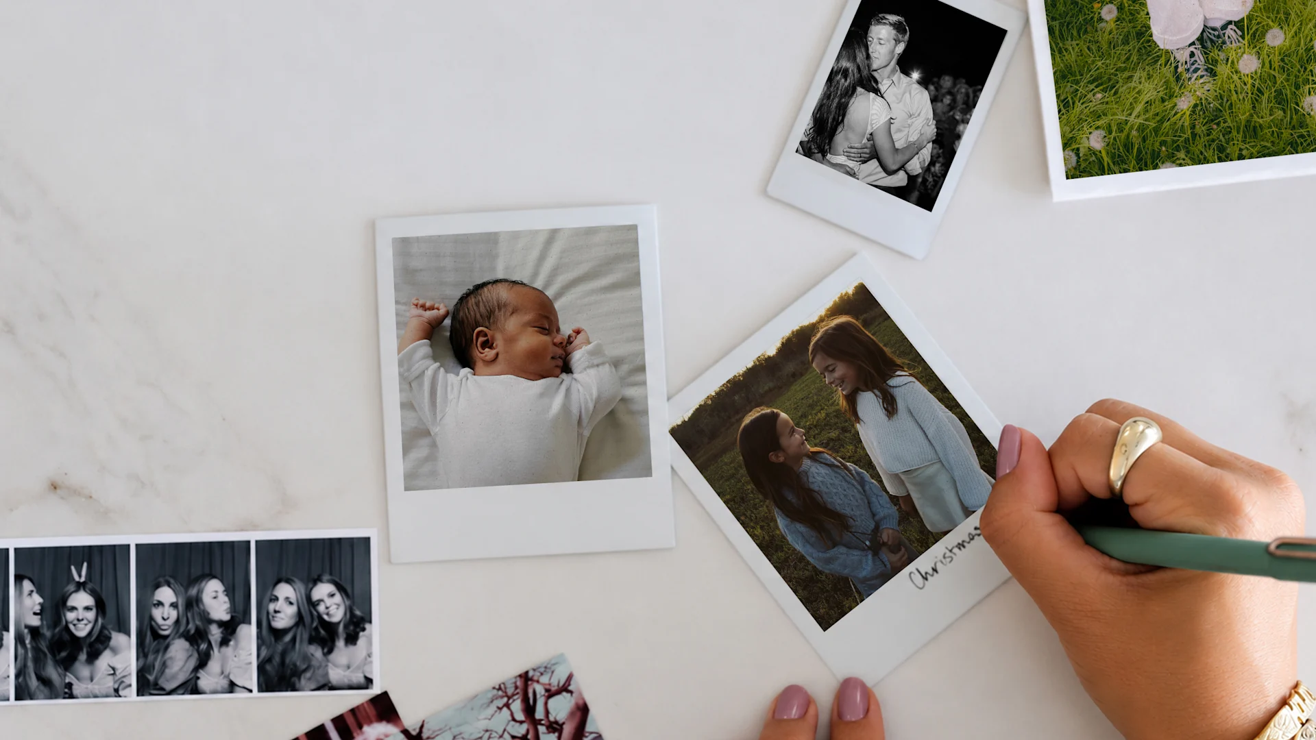 A hand writing “Christmas” on an instant-style photo of two girls, among scattered photos of a newborn, a couple, and a photo strip