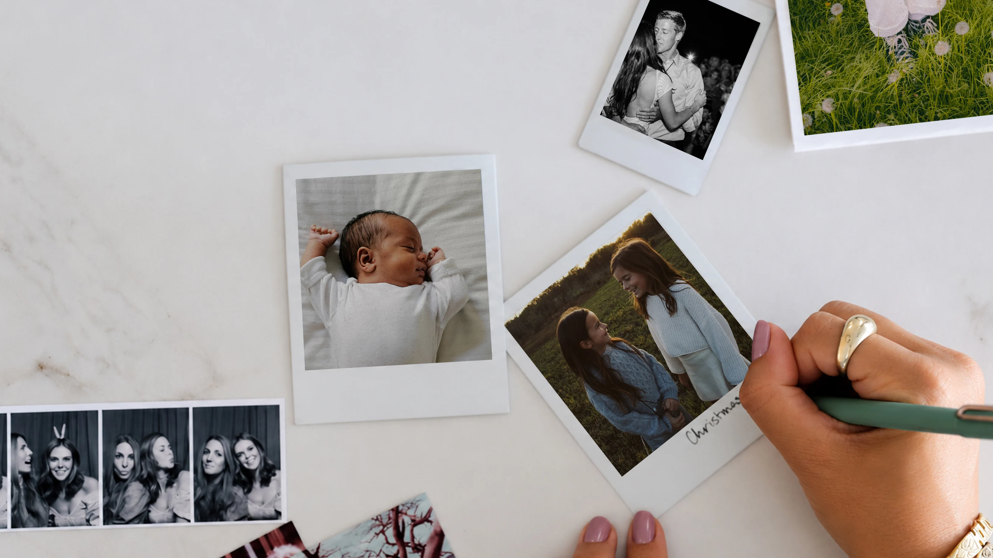 A hand writing “Christmas” on an instant-style photo of two girls, among scattered photos of a newborn, a couple, and a photo strip