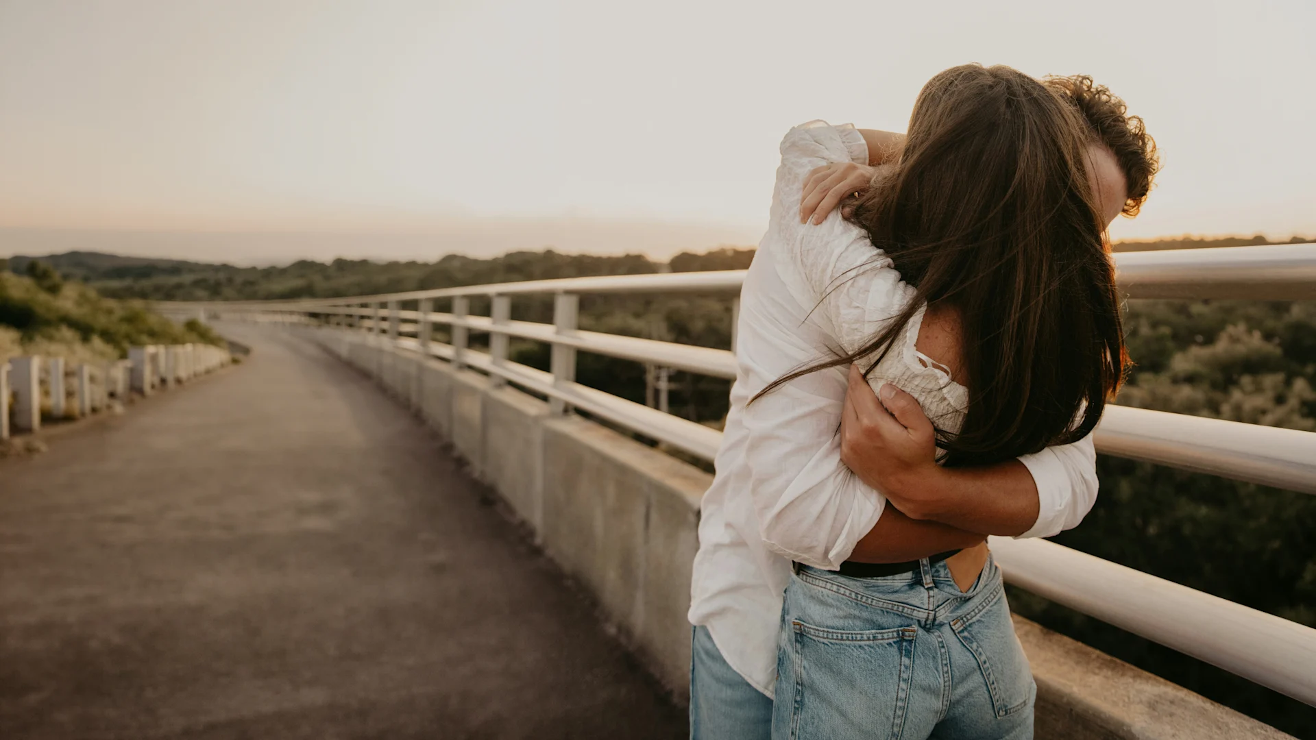 Couple sharing a warm embrace on a quiet bridge at sunset, surrounded by a peaceful natural landscape.
