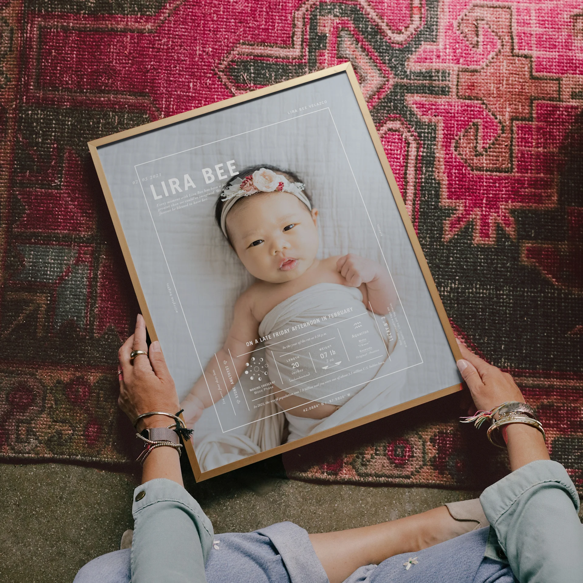 A framed Birth Stat Print featuring a newborn photograph is held over a patterned rug.