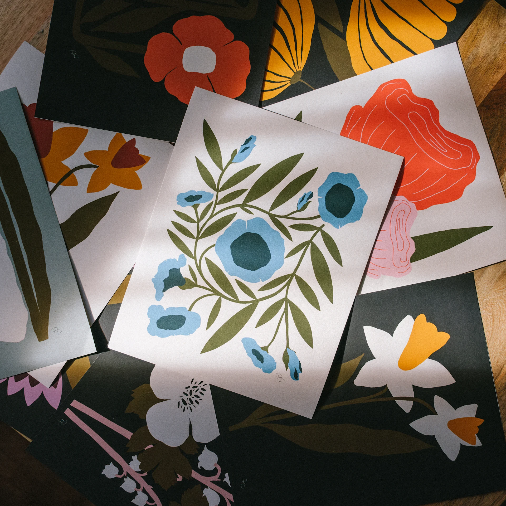 Colorful botanical art prints by McKenzie Parrott spread across a wooden table, with blue, orange, and yellow florals.