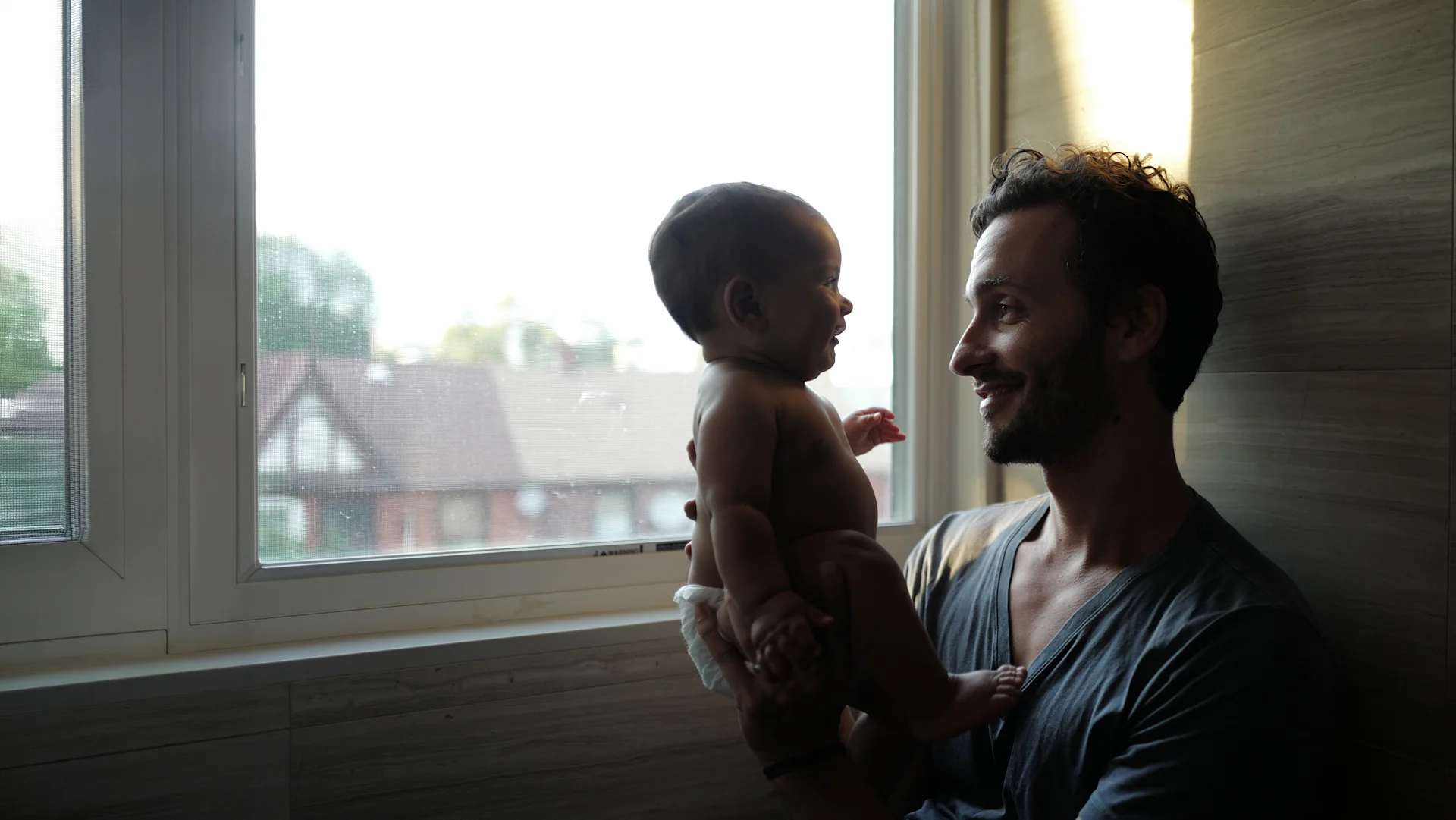 Warm, candid photograph of a smiling father in a gray T-shirt holding his baby in a white diaper beside a sunlit window. They gaze at each other, with soft natural light illuminating their faces and rooftops visible outside.