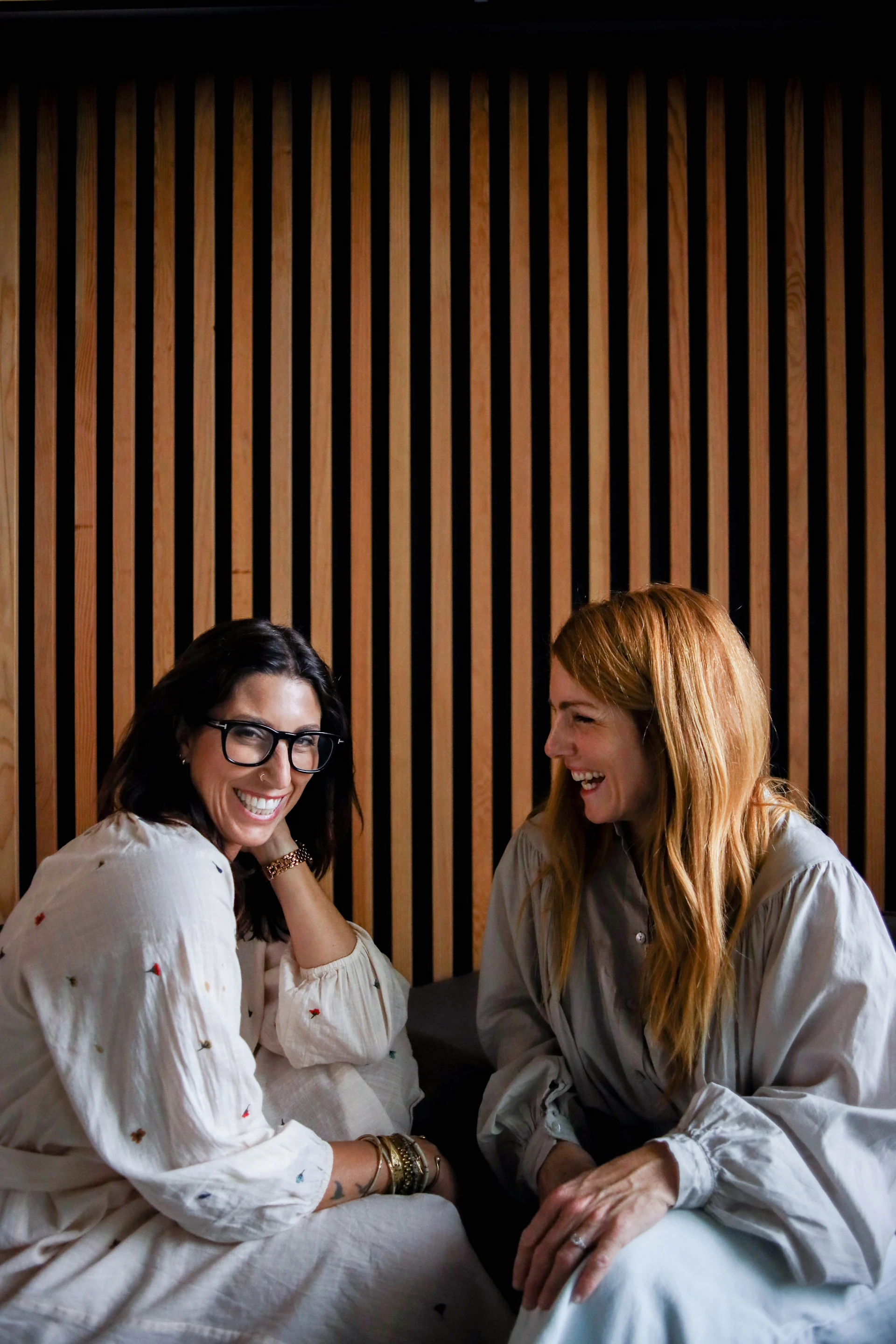 Candid portrait of Still Novel’s two female founders laughing together in front of a vertical wood-paneled wall. One has dark hair, black glasses, and a white embroidered dress; the other has long strawberry-blonde hair and a light blue blouse.