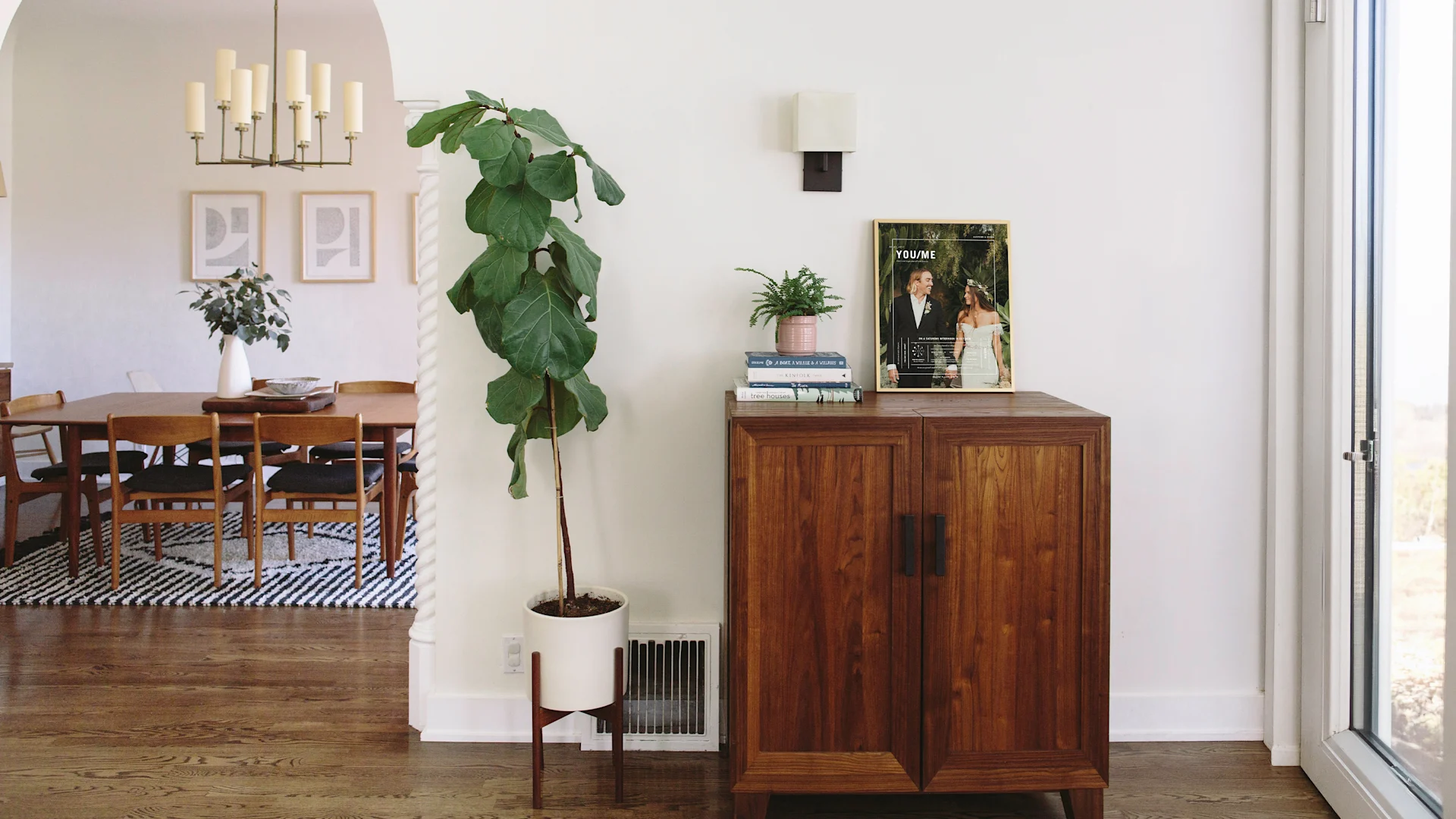 Framed wedding print displayed on a wooden cabinet beside books and a potted plant in a bright dining space.