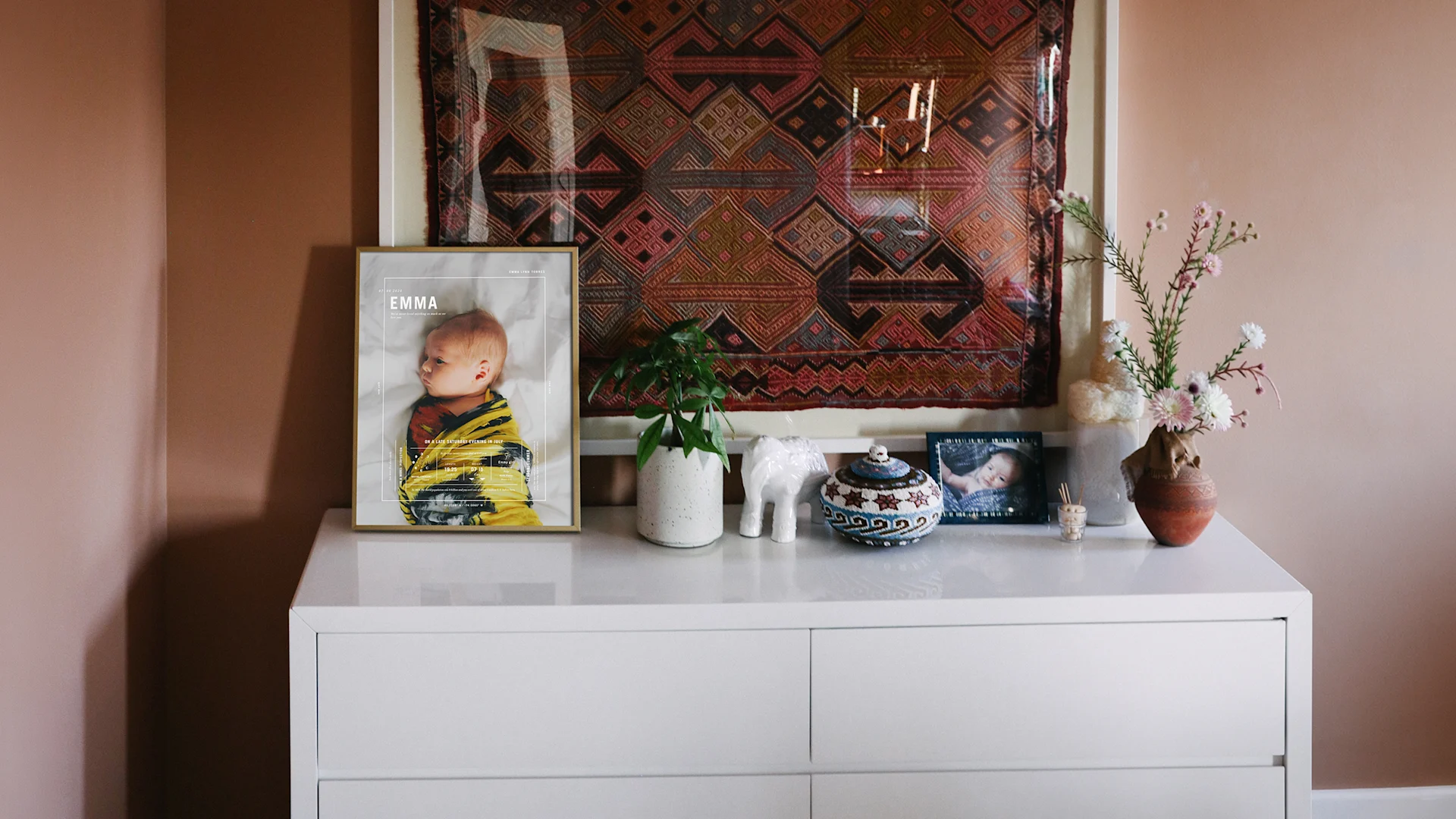 Framed birth stat print displayed on a white dresser beside a plant and ceramics, beneath a patterned textile.