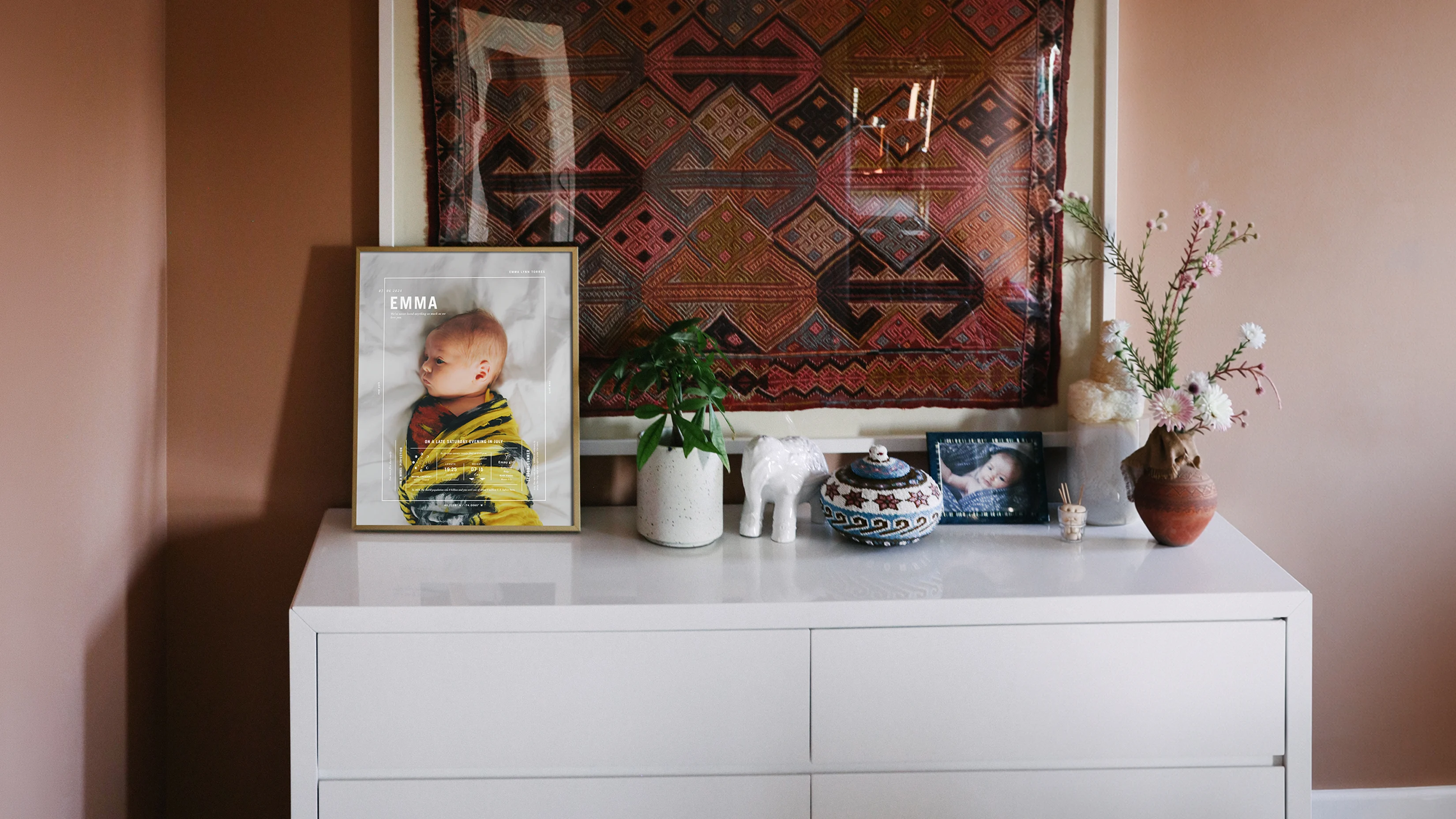 Framed birth stat print displayed on a white dresser beside a plant and ceramics, beneath a patterned textile.