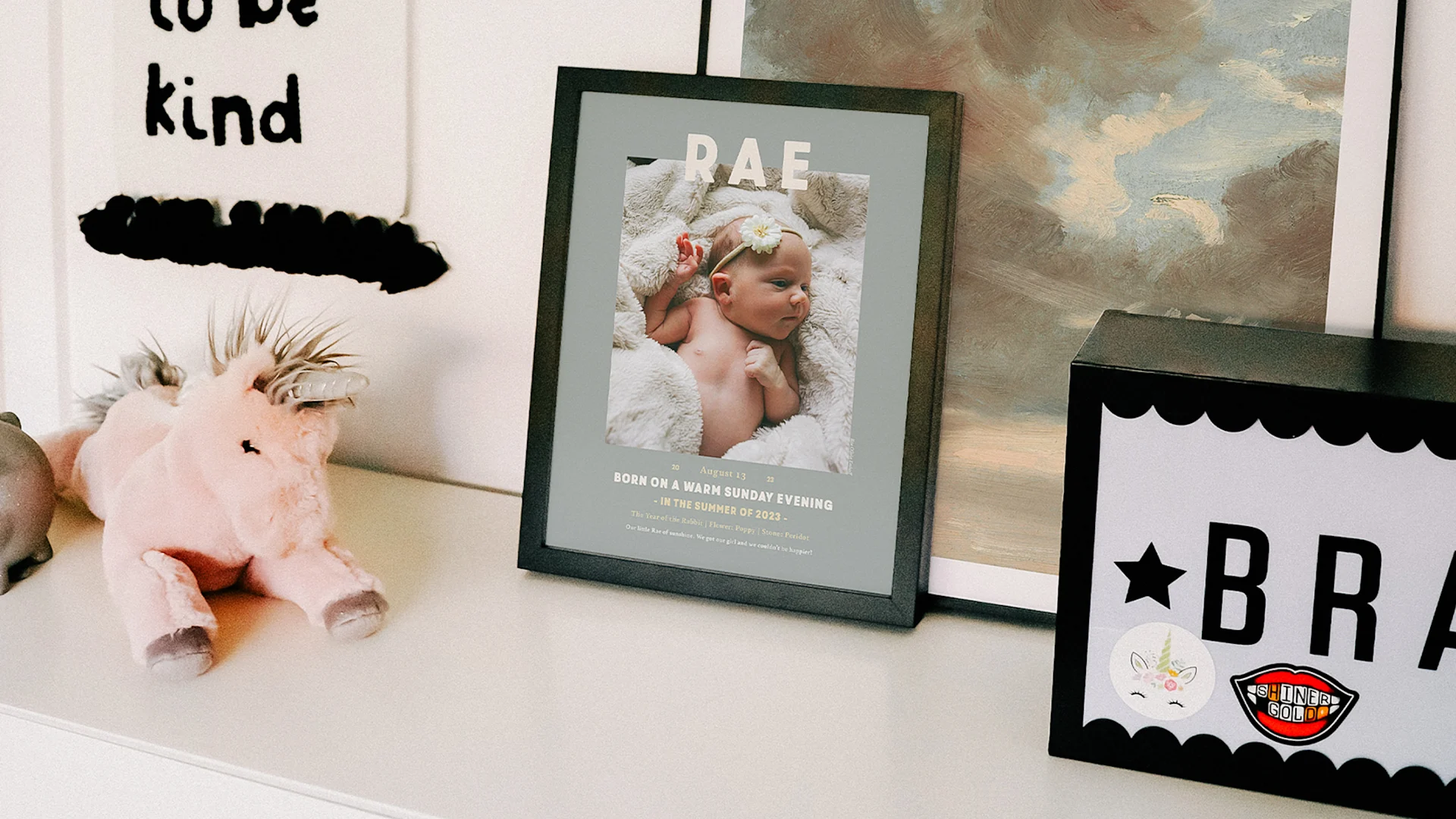 A green-framed Rae Birth Stat Snapshot Print rests on a nursery dresser beside a unicorn toy and other framed artwork.