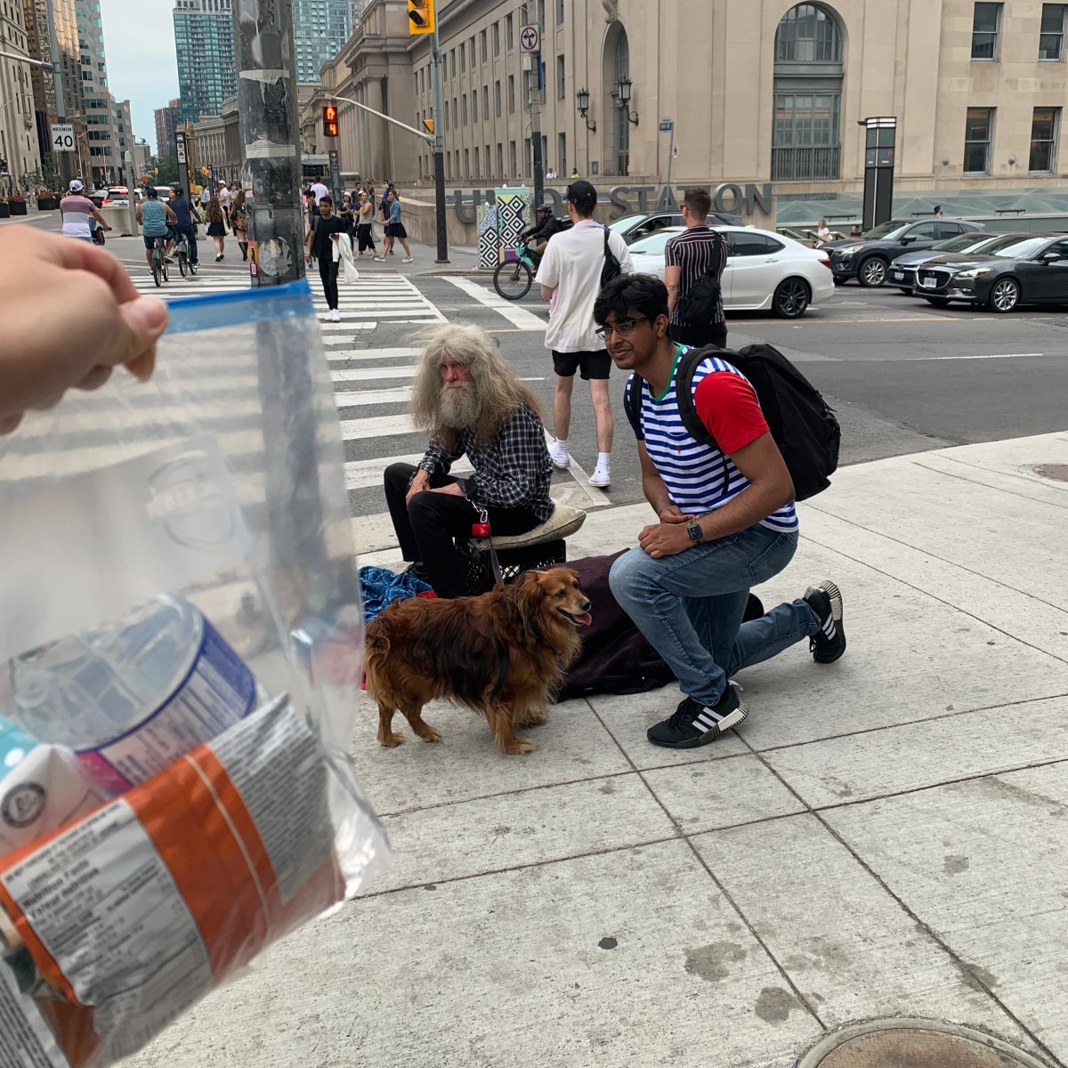 A homeless individual and their dog are seen at a street corner in front of Toronto Union Station. A member of Blankets for T.O. is seen kneeling next to him.