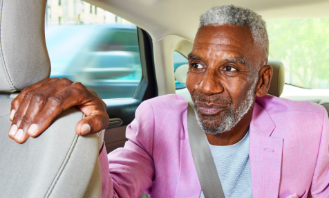 man with graying hair and beard in back seat with seat belt on placing hand on front seat