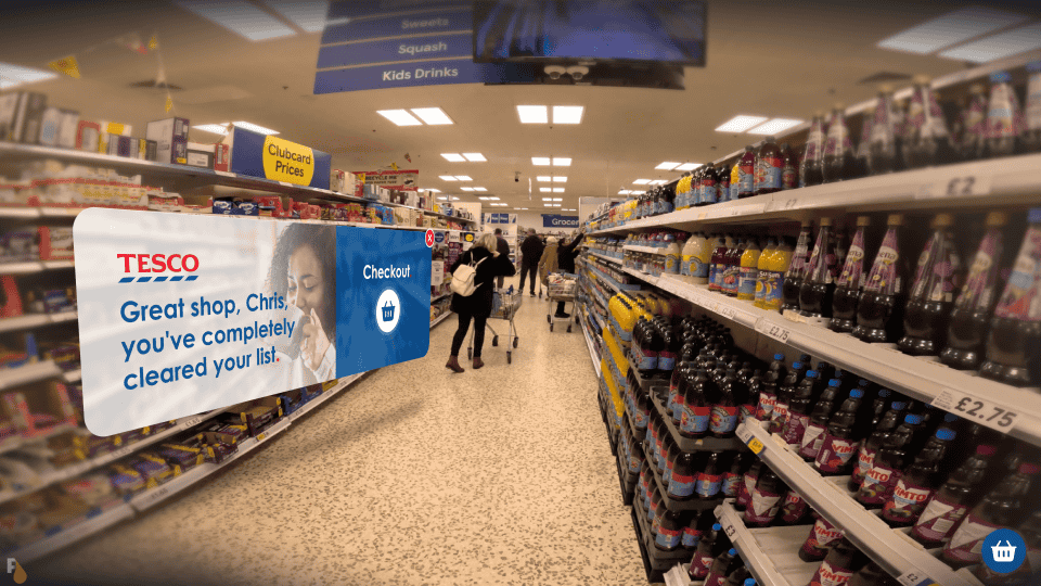 A first-person view of a grocery store aisle, with an augmented reality banner from Apple Vision Pro AR glasses displaying 'TESCO Great shop, Chris, you've completely cleared your list.' and a checkout icon, indicating the completion of the shopping list.