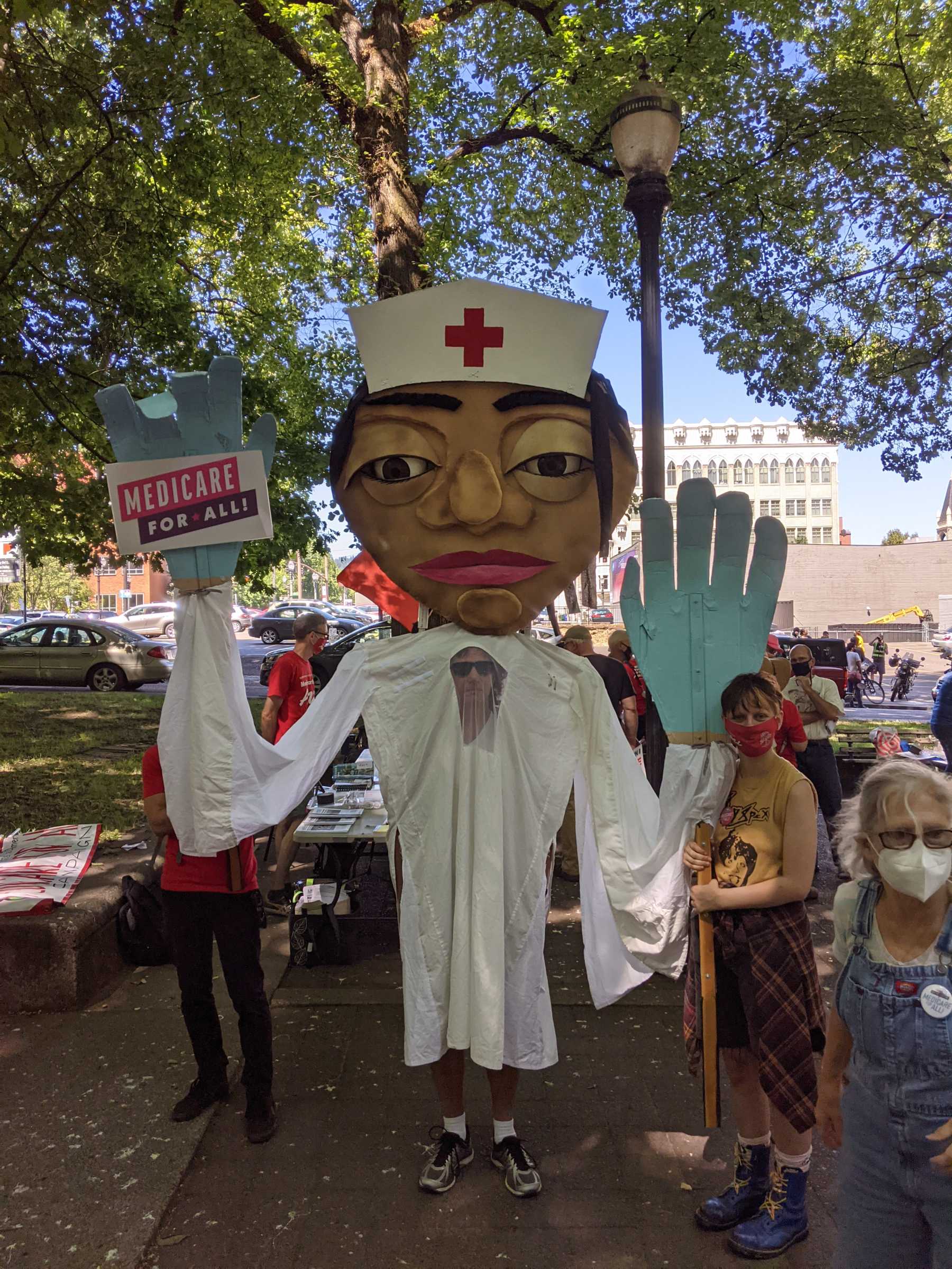 A person wears a big nurse puppet. In one hand, the nurse holds a sign for Medicare for All.