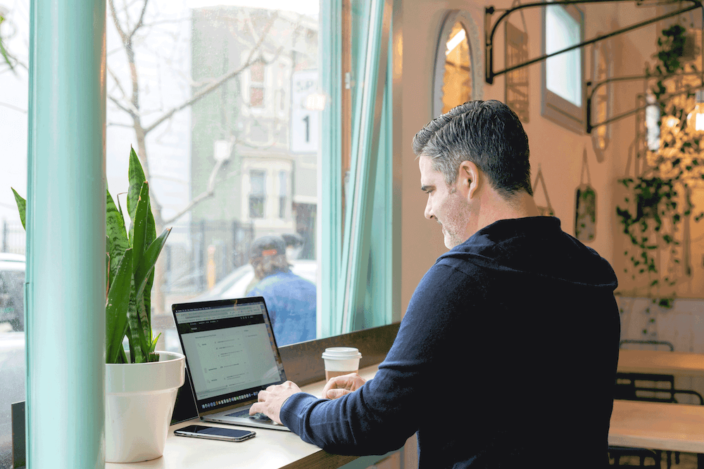 A man in a cafe window working on a laptop with a cup of coffee next to him.