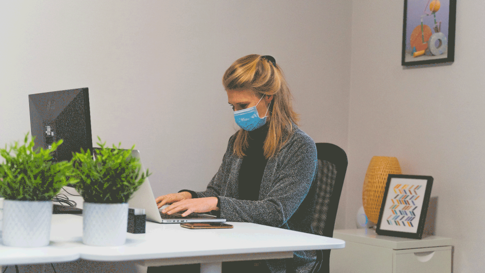 A blonde woman wearing a blue surgical mask is working on a laptop at a white desk set up with a green potted plant.