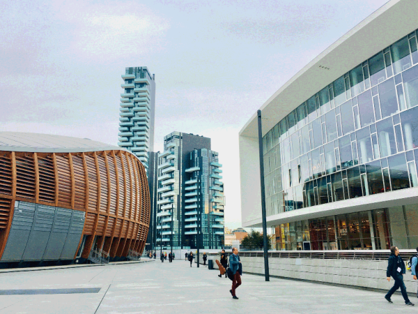 One rounded building with a wooden frame next to two taller skyscrapers and across from a building with a glass front. People are walking in the middle of the buildings.