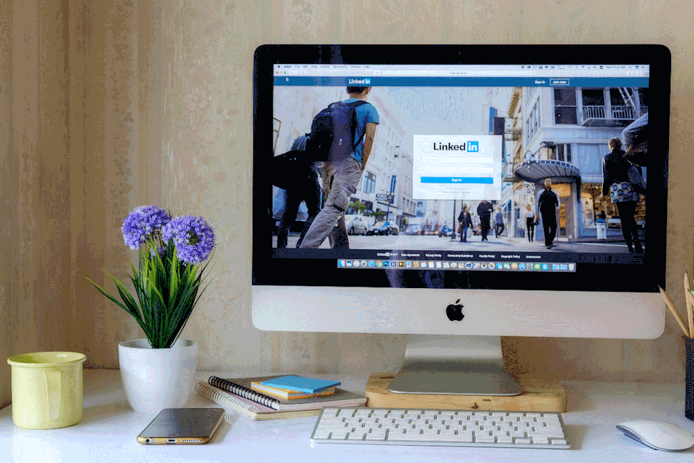 LinkedIn homepage displayed on Apply computer monitor with a tan wall background. White desk includes yellow pot, white pot with purple flowers, an assortment of notebooks, a Bluetooth keyboard, and a wireless mouse.