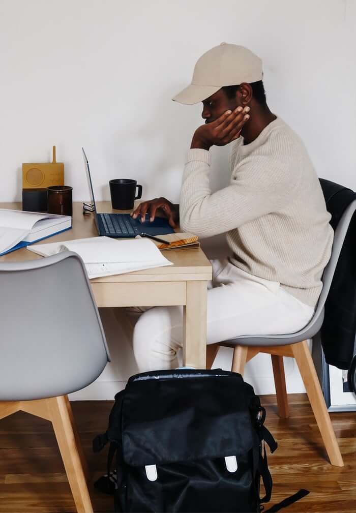 Young man working at laptop. He's wearing a beige hat and sweater.