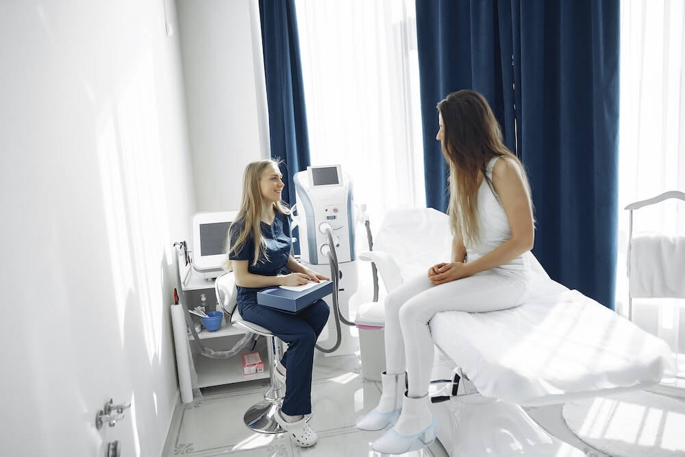 A white long blonde haired nurse working with a patient sitting on a cot in an exam room.