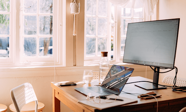 A desk with a laptop on it connected to an external monitor. A glass of water and computer accessories are also on the desk. An empty chair is slightly pulled out next to the desk.