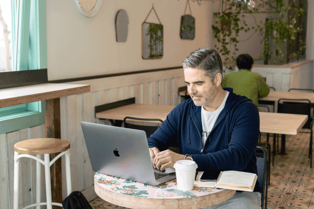 Man in a cafe working on Apple laptop with book and coffee cup on the table next to him