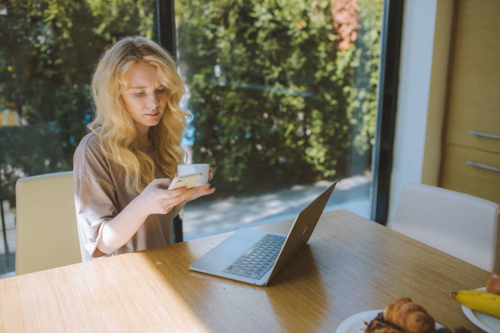 Blonde woman looking at phone with open laptop in front of her. Large glass window behind her shows bushes.