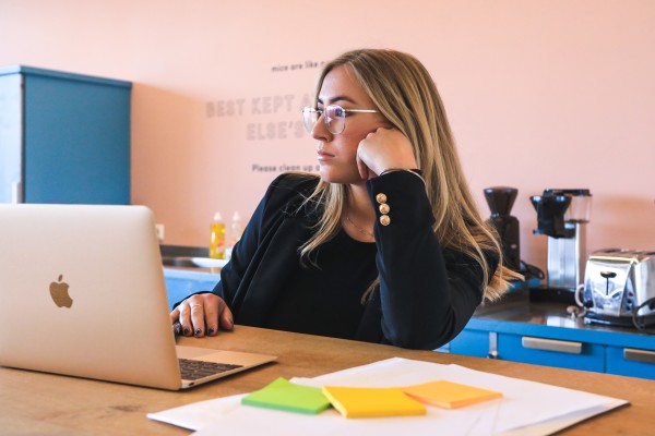 Woman in long sleeved black shirt sitting at a table with a MacBook. She has blonde hair and glasses. Foreground has post-it note packets and paper. Background haws a blue counter with coffee maker and toaster.