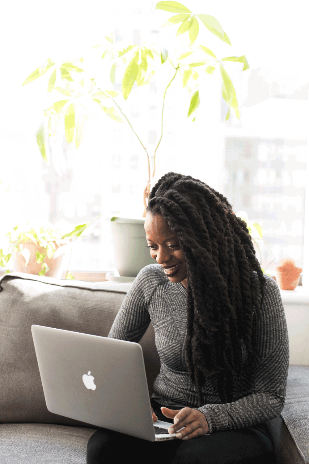A woman with long dreadlocks sitting on a grey couch smiling while using her Apple laptop. 