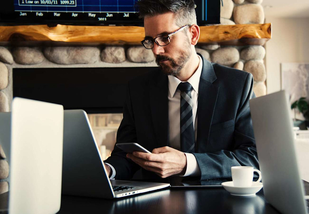 Man in suit and tie holding phone while looking at laptop. Two other laptops are placed in the foreground, implying he is at a shared working space or coffee shop.