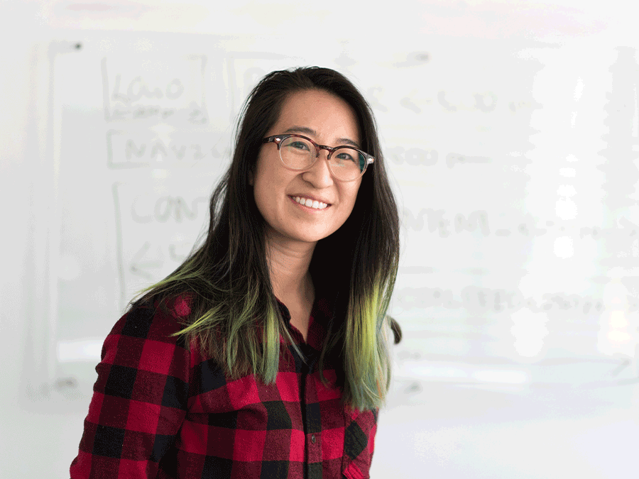 Young woman teaching in glasses and red and black plaid shirt smiling. There is a whiteboard in the background.