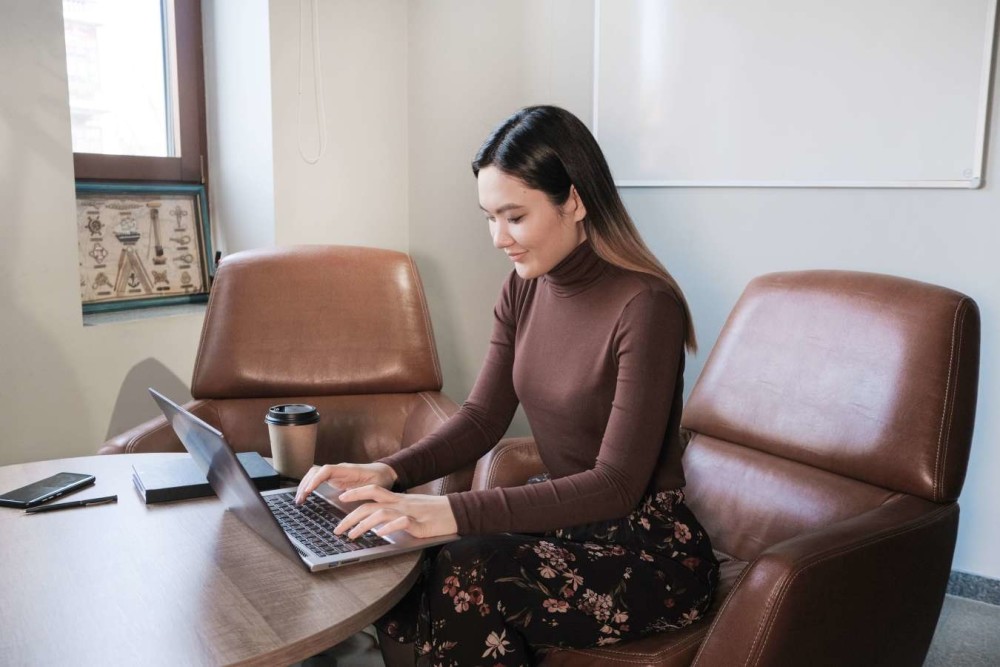 Woman on a leather chair on a laptop computer in a conference room setting. She's wearing a brown turtleneck and a floral skirt.