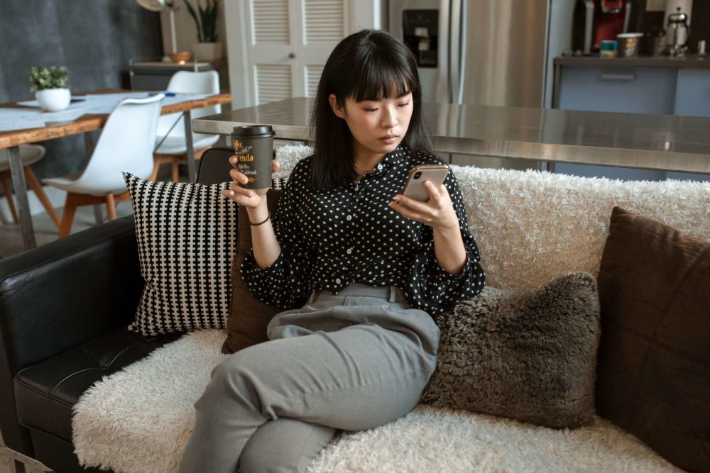 A woman seated on the couch while looking at her phone and holding a coffee cup. 