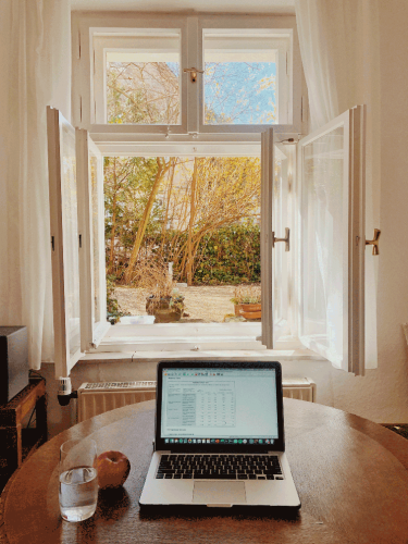 A table with an open laptop, an apple, and a glass of water sits in front of an open window showing trees and a hedge.