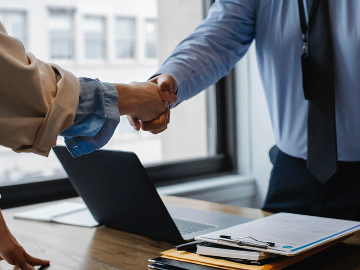 Two people shaking hands with open laptop and notepads on table