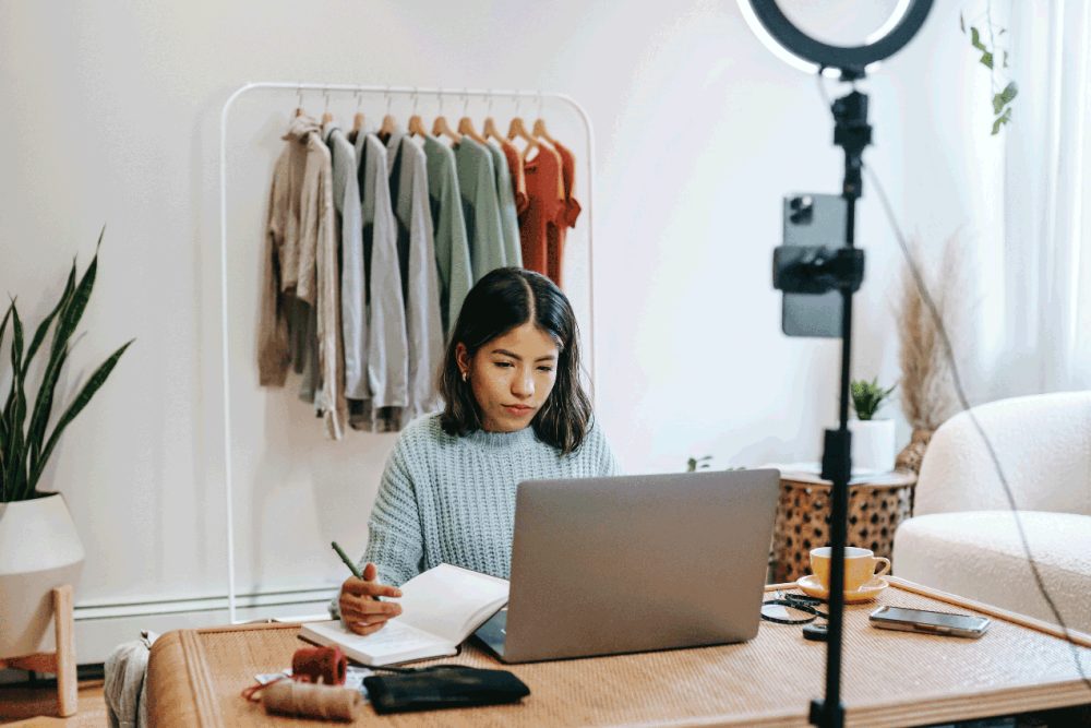 A woman in a light blue sweater works at a desk on her laptop while she takes notes in her notebook. Behind her is a garment rack full of different clothes. In front of her is a ring light.