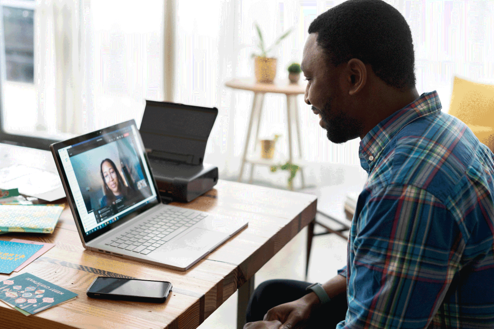 A black man in a plaid shirt is smiling at a laptop computer, on which he is videoconferencing with a woman. The computer is on top of a wooden table, and a cell phone is next to the computer.