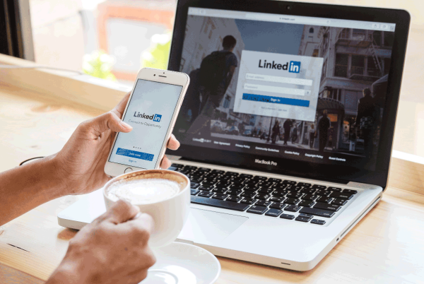 The LinkedIn login screen is on a laptop, and a pair of hands, one holding a mobile phone and the other holding a ceramic mug full of coffee, are in front of the laptop. 