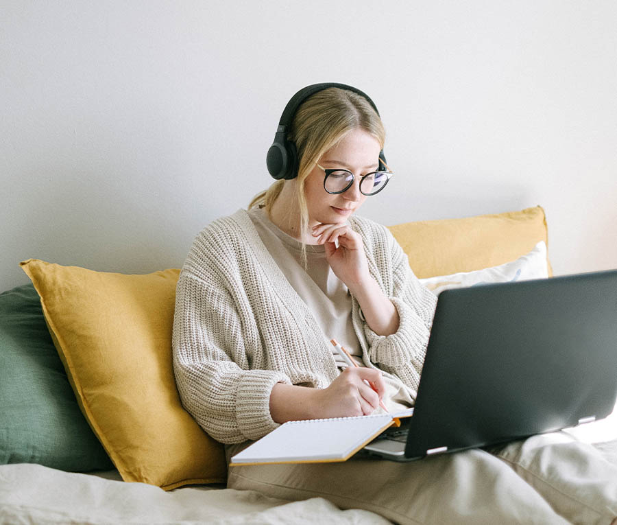 Woman with headphones and glasses sitting in bed with a notebook and laptop on her lap