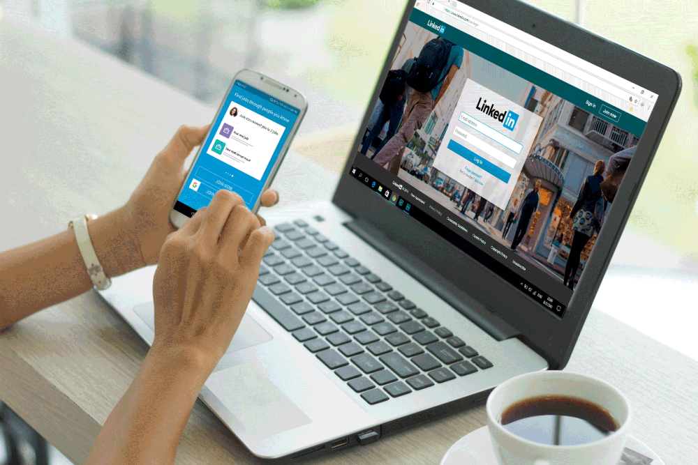 Woman holds phone with LinkedIn app displayed while a laptop also shows LinkedIn homepage. The thin table hold laptop also has a cup of coffee in a white mug with a saucer.