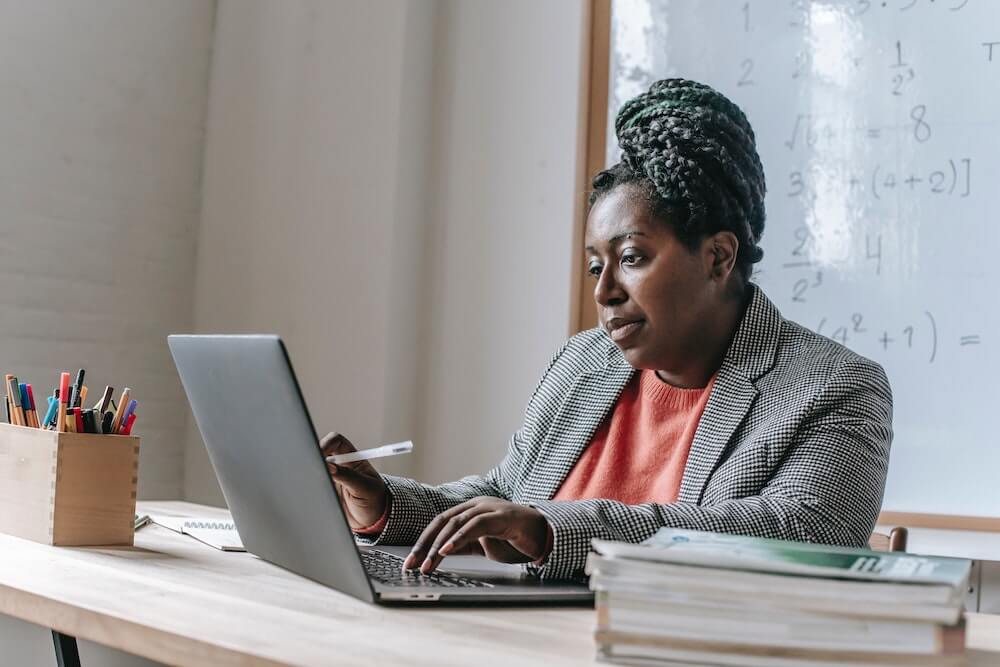 A Black woman wearing a blazer and coral shirt working at a desk at a laptop.