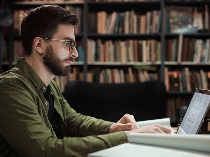 Man with glasses and beard reading a laptop in a library