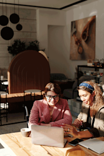 Two women sitting at a wooden table looking together at a laptop screen.