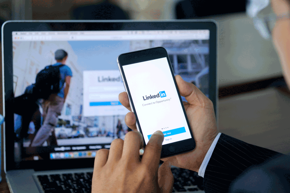 Man with glasses looks at LinkedIn app on a phone while the LinkedIn site is open on a laptop screen in the background. His finger is tapping the blue login icon.