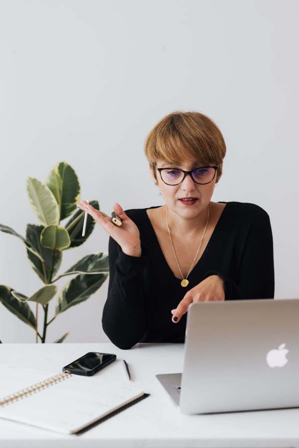 Woman talking to open Apple computer, gesturing with her hand as though explaining or engaged in conversation. She has short hair, glasses, a black top, and a plant in the background.