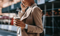A woman wearking a khaki trenchcoat walking on a city street, talking on the phone and holding a paper cup of coffee.