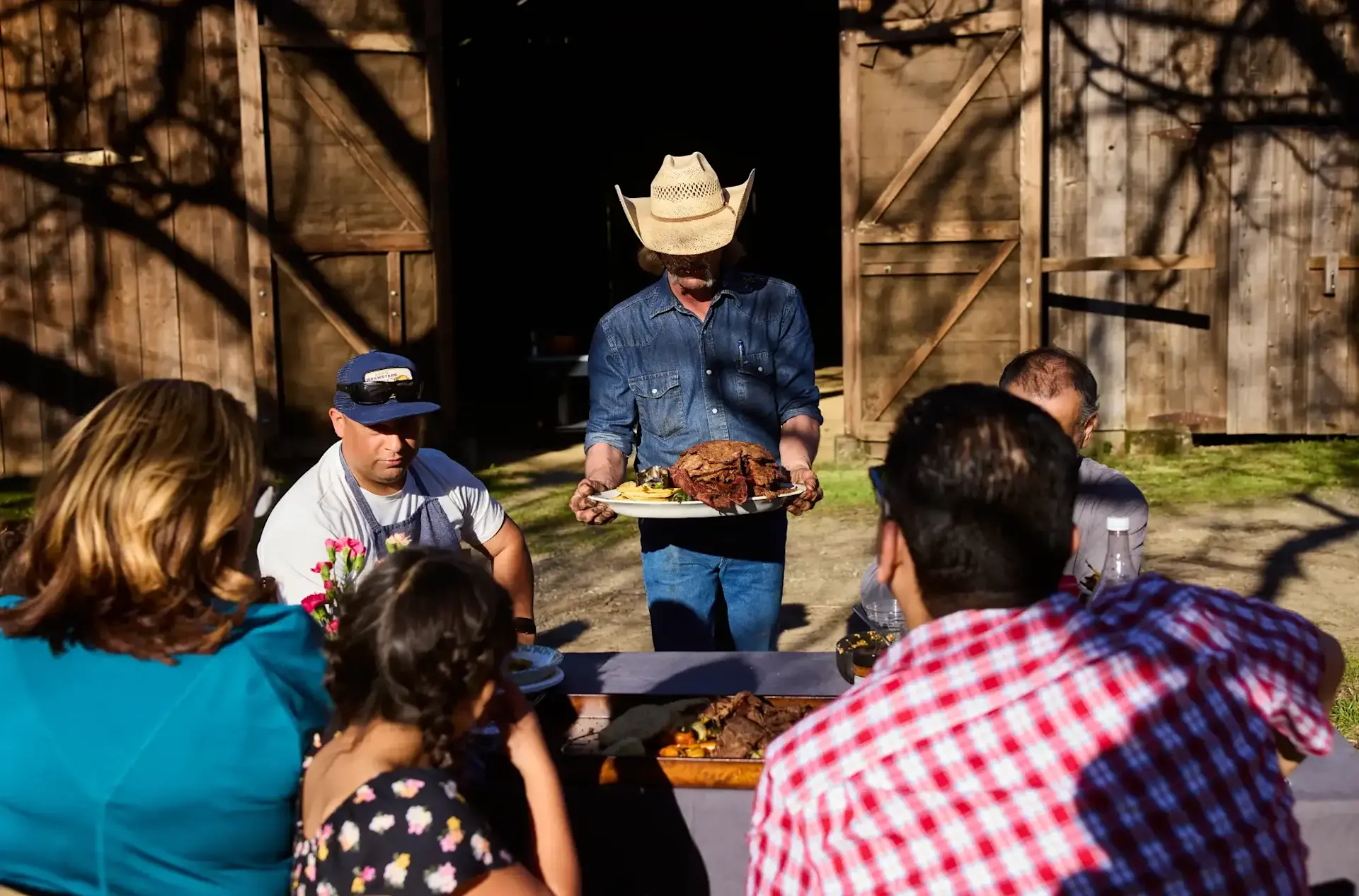 A man in a cowboy hat with a tray of steak serving it to a family sitting at a picnic table