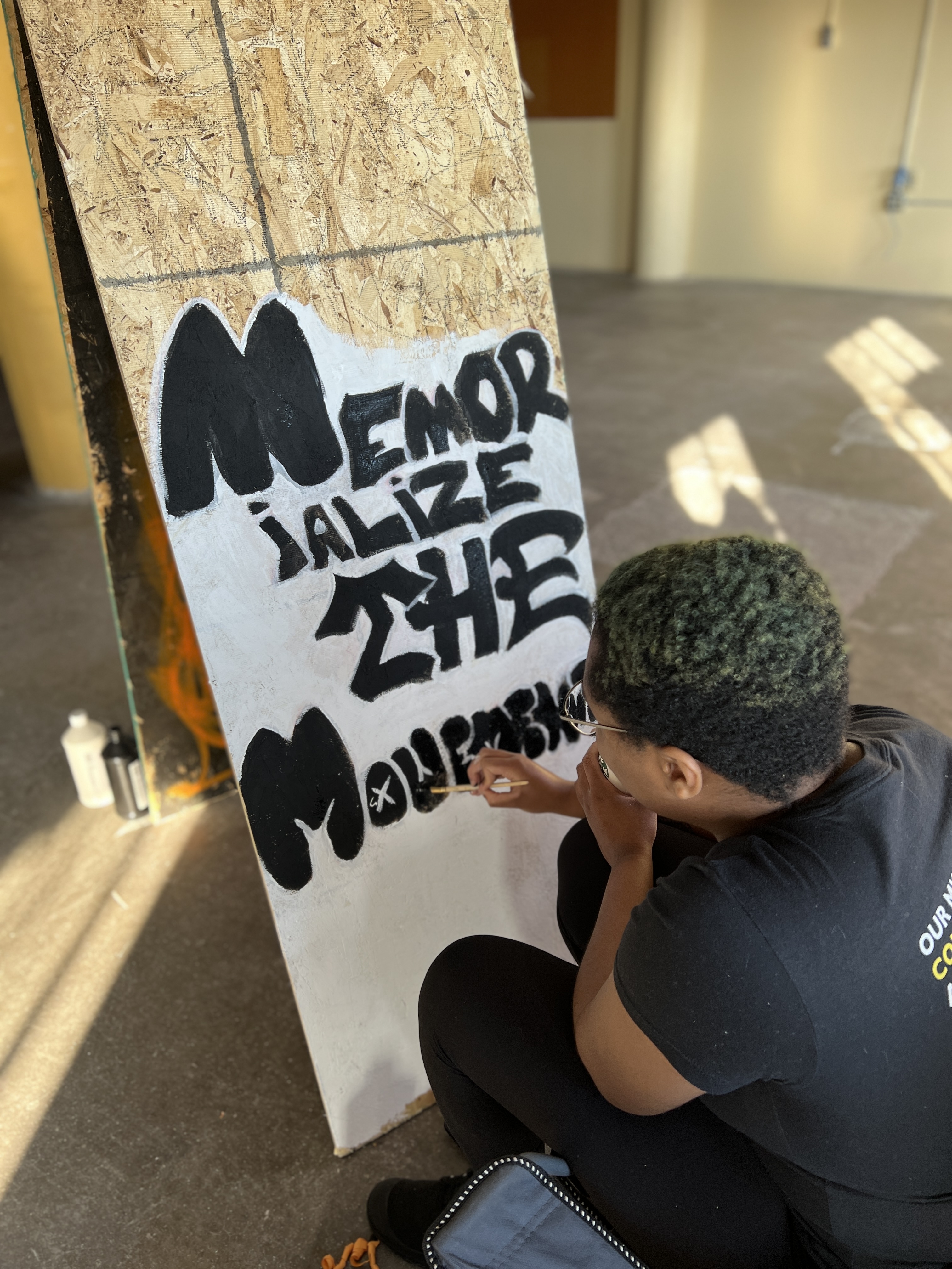 A person painting a piece of plywood with the words "Memorialize the Movement"